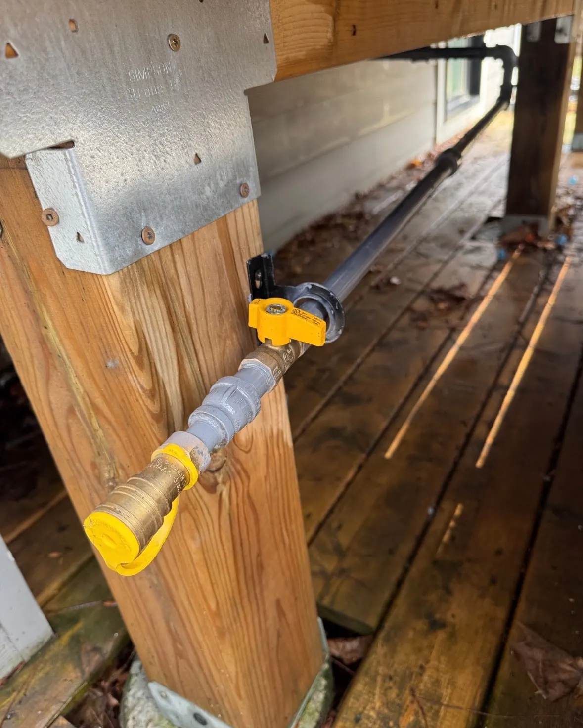 Natural gas line under a wooden deck ending in a yellow shut-off valve and quick-connect fitting for a barbecue